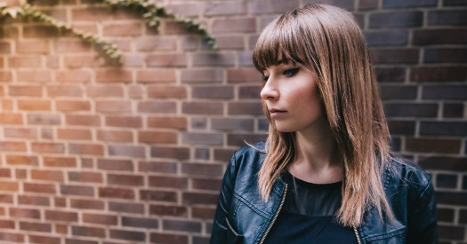 Brunette woman wearing a black jacket looks down anxiously while standing in front of a red brick wall