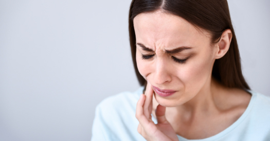 Brunette woman cringes and touches her cheek due to tooth pain from an untreated cavity