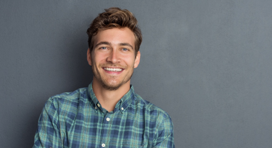 Brunette man wearing a plaid shirt smiles against a gray wall after working hard to reverse his gum disease